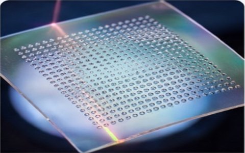 Close-up of a transparent chip with a grid of tiny circular holes reflecting colorful light at the edges.