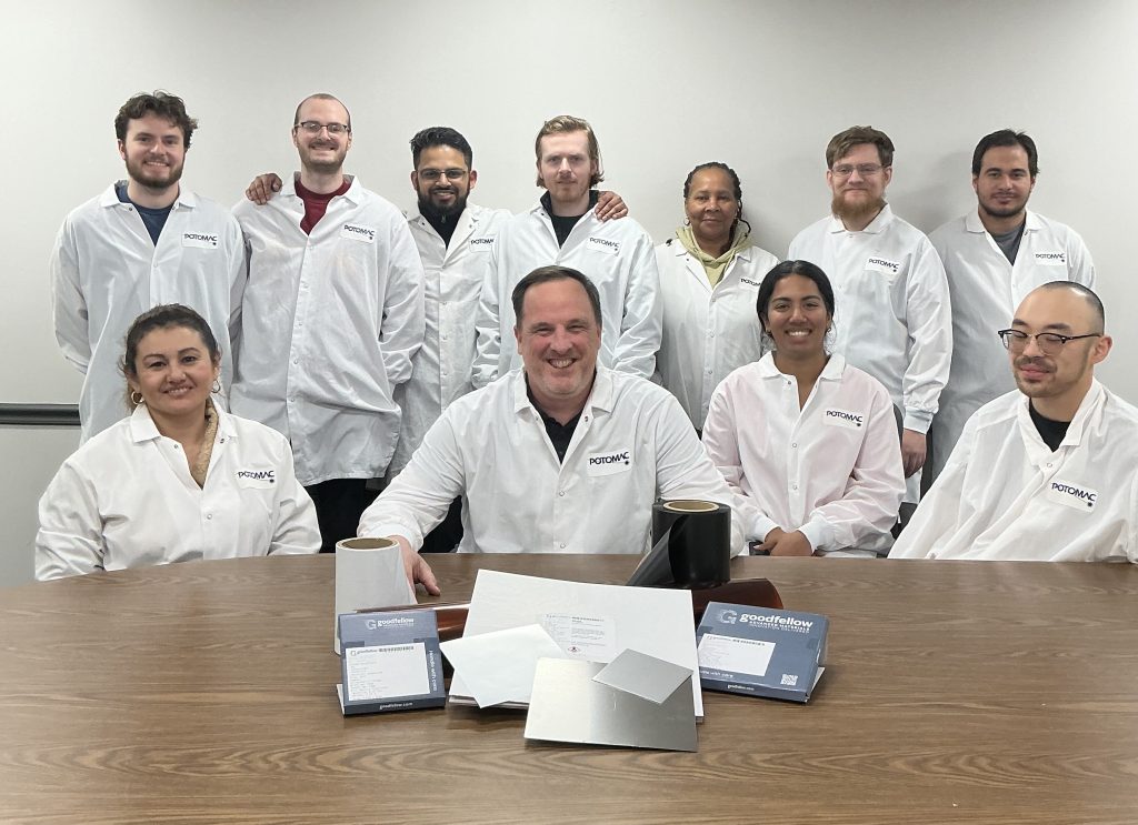 Group of twelve people in white lab coats posing for a team photo around a wooden table, in a bright room.